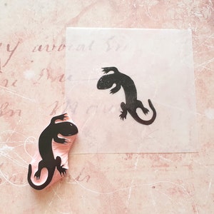 May include: A black lizard stamp and its impression on a piece of paper. The stamp is mounted on a pink base. The paper with the lizard impression is on a pink background with script.