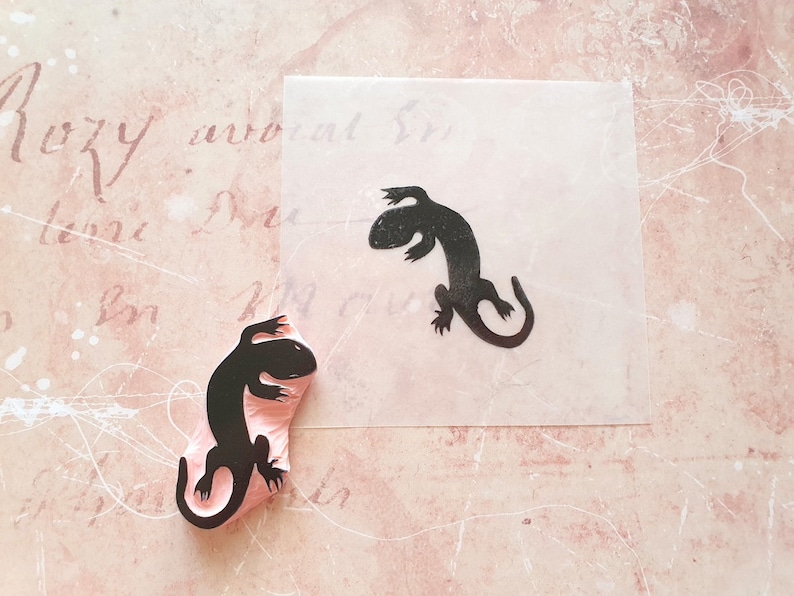 May include: A black lizard stamp and its impression on a piece of paper. The stamp is mounted on a pink base. The paper with the lizard impression is on a pink background with script.