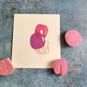 May include: A cream-colored card with a pink and purple abstract print, accompanied by three pink rubber stamps. The stamps have various shapes, including a rounded rectangle, a U-shape, and a textured square. The background is a mottled blue-gray.
