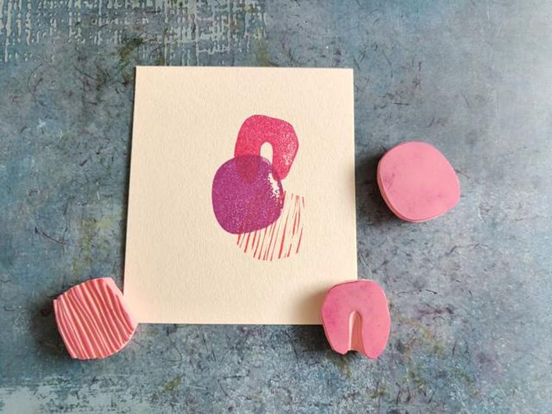 May include: A cream-colored card with a pink and purple abstract print, accompanied by three pink rubber stamps. The stamps have various shapes, including a rounded rectangle, a U-shape, and a textured square. The background is a mottled blue-gray.