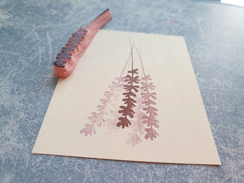 May include: A wooden stamp with a design of a bunch of lavender flowers. The stamp is pink and the design is brown. The stamp is on a piece of paper with a pink impression of the design.