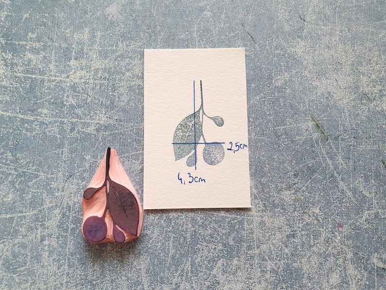 以下が含まれることがあります： 葉と3つのベリーのデザインが施された木製のスタンプ。スタンプの幅は4.3cm、高さは2.5cmです。