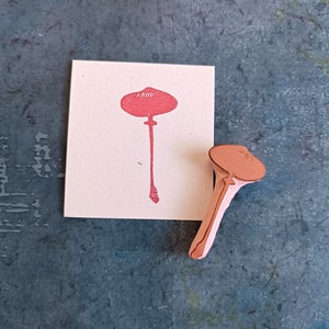 May include: A wooden mushroom-shaped stamp with a pink rubber design of a mushroom. The stamp is on a white card with a pink mushroom design.