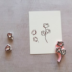 May include: A floral stamp with a pink handle and black design, alongside a matching ink drawing on white paper. Two additional stamps are also visible. The design features stylized flower buds and stems.