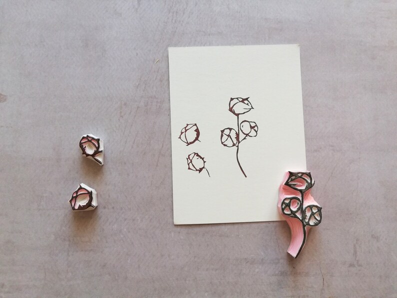 May include: A floral stamp with a pink handle and black design, alongside a matching ink drawing on white paper. Two additional stamps are also visible. The design features stylized flower buds and stems.