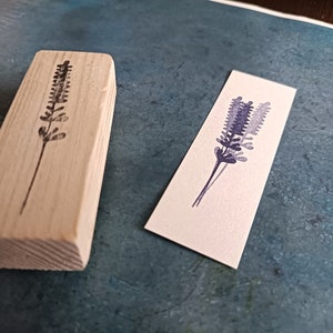 May include: A wooden block stamp with a black outline of a lavender flower. The stamp is next to a white card with a purple watercolor lavender flower.