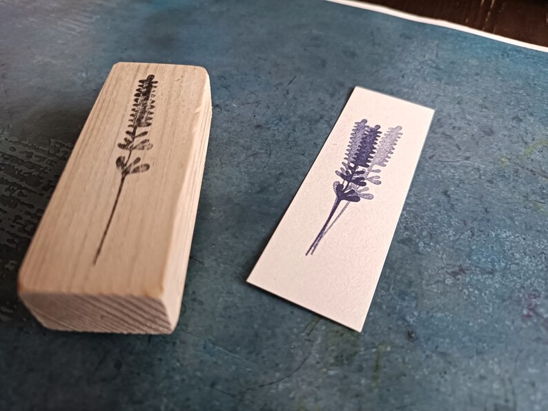 May include: A wooden block stamp with a black outline of a lavender flower. The stamp is next to a white card with a purple watercolor lavender flower.