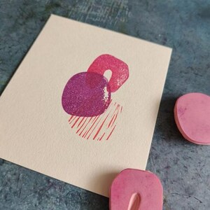 May include: A cream-colored card features a print of abstract shapes in shades of pink and purple. The design includes a circle, a U-shape, and a striped rectangle. Two pink rubber stamps are visible.