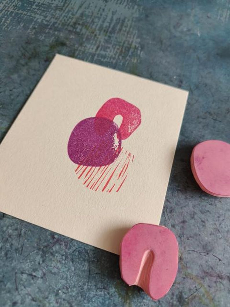 May include: A cream-colored card features a print of abstract shapes in shades of pink and purple. The design includes a circle, a U-shape, and a striped rectangle. Two pink rubber stamps are visible.