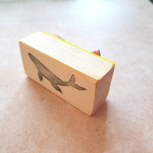 May include: A wooden stamp with a yellow edge, featuring a black silhouette of a whale. The stamp is designed for crafting and creating unique designs.