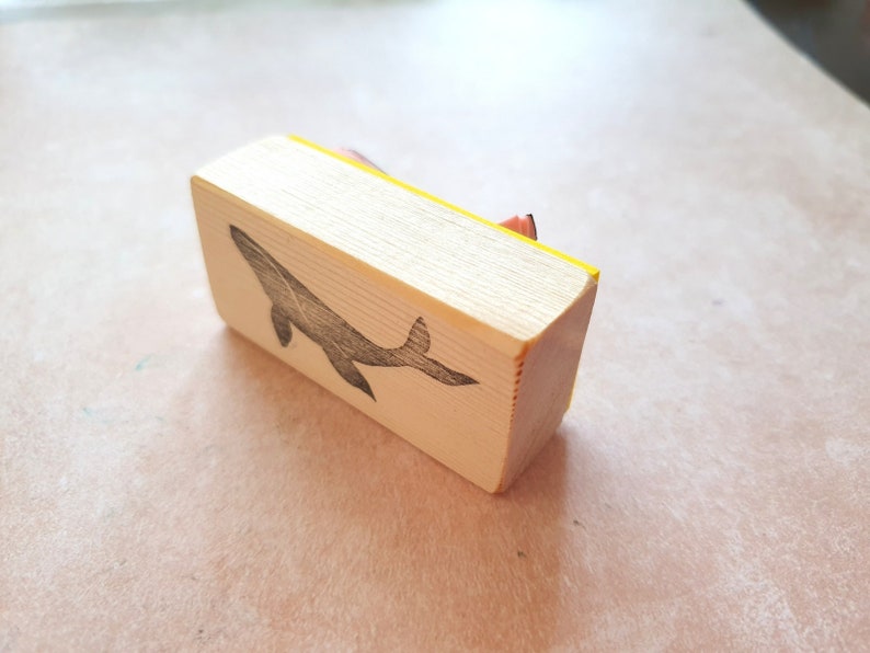 May include: A wooden stamp with a yellow edge, featuring a black silhouette of a whale. The stamp is designed for crafting and creating unique designs.