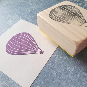 Pode incluir: Um carimbo de madeira com um desenho de bal&atilde;o de ar quente roxo. O carimbo est&aacute; em um cart&atilde;o branco com uma impress&atilde;o de bal&atilde;o de ar quente roxo.