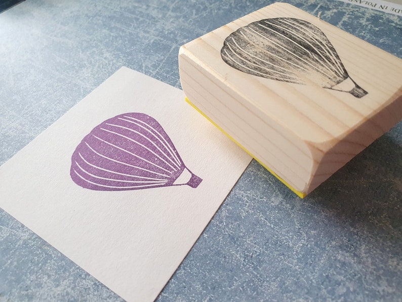 Pode incluir: Um carimbo de madeira com um desenho de bal&atilde;o de ar quente roxo. O carimbo est&aacute; em um cart&atilde;o branco com uma impress&atilde;o de bal&atilde;o de ar quente roxo.
