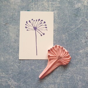 May include: A pink rubber stamp with a detailed dandelion design. The stamp is on a blue textured surface next to a card with a purple dandelion print.