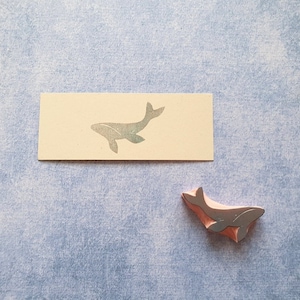 May include: A close-up of a whale-shaped rubber stamp. The stamp is a light brown color and has a silver whale silhouette. The stamp is sitting on a light blue surface.