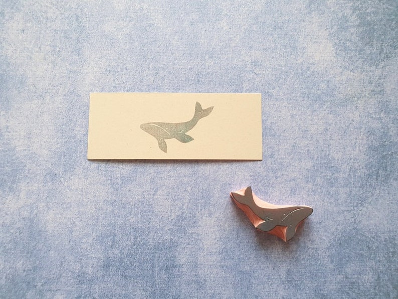 May include: A close-up of a whale-shaped rubber stamp. The stamp is a light brown color and has a silver whale silhouette. The stamp is sitting on a light blue surface.