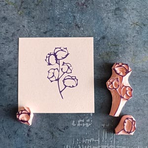 May include: A cotton plant stamp with a floral design is shown next to a stamped image of the same design on a square piece of paper. The stamp is made of pink rubber with a dark purple floral design.