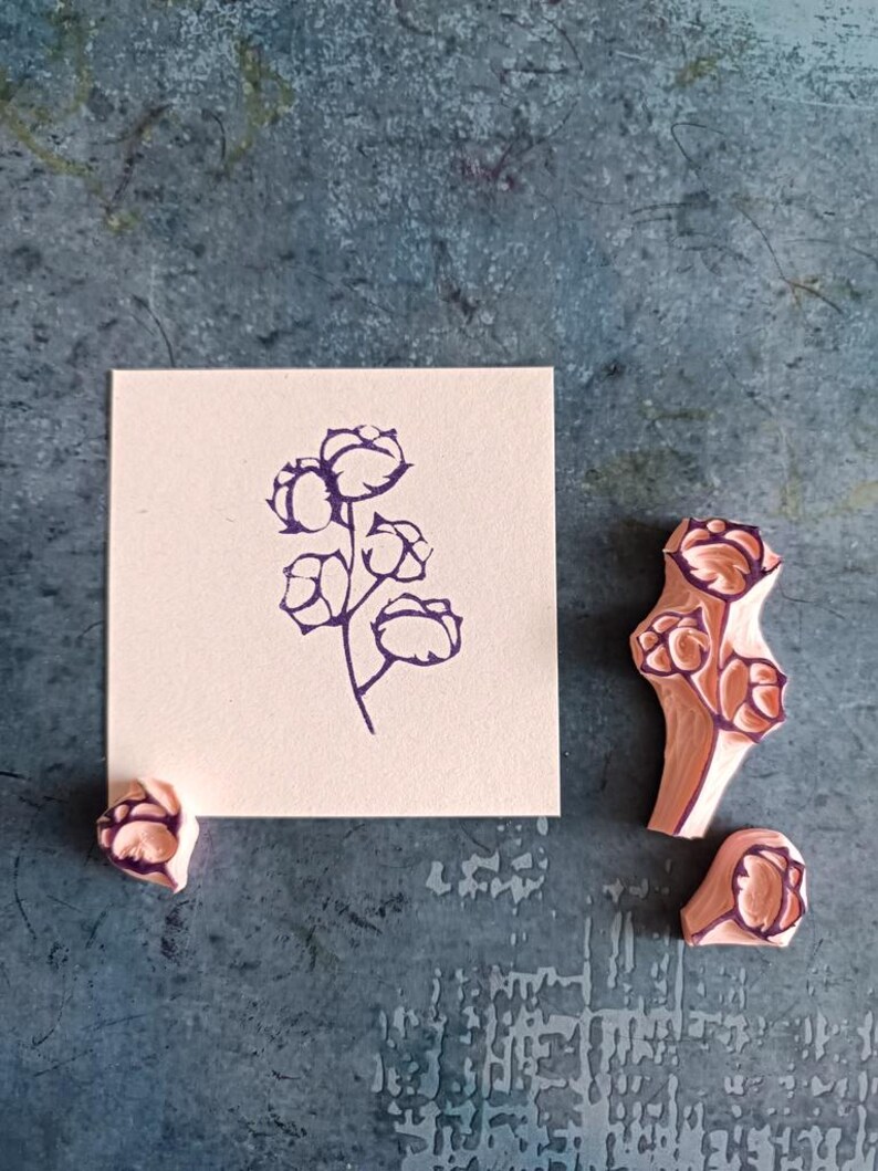 May include: A cotton plant stamp with a floral design is shown next to a stamped image of the same design on a square piece of paper. The stamp is made of pink rubber with a dark purple floral design.