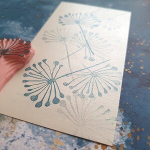 May include: A close-up of a rubber stamp with a dandelion design. The stamp is pink and has a black outline. The dandelion design is in a light blue color and has been stamped on a white card.