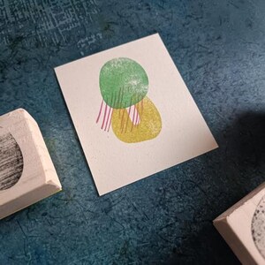 May include: A small, off-white card with a green and yellow abstract design, with pink and white stripes. Three wooden stamps with black designs are scattered around the card on a textured blue surface.