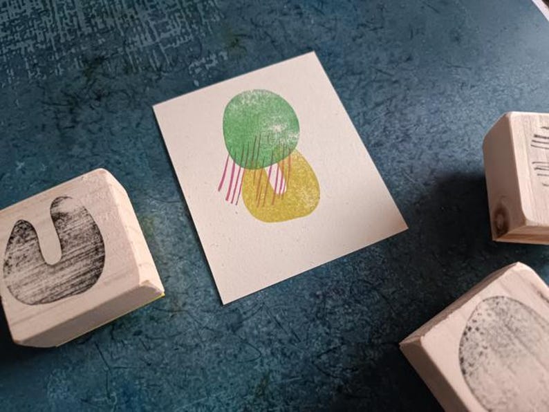 May include: A small, off-white card with a green and yellow abstract design, with pink and white stripes. Three wooden stamps with black designs are scattered around the card on a textured blue surface.