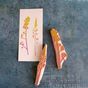 May include: Two pink rubber stamps with a yellow and orange floral design. The stamps are on a blue textured surface. A card with a yellow and orange floral design is in the background.