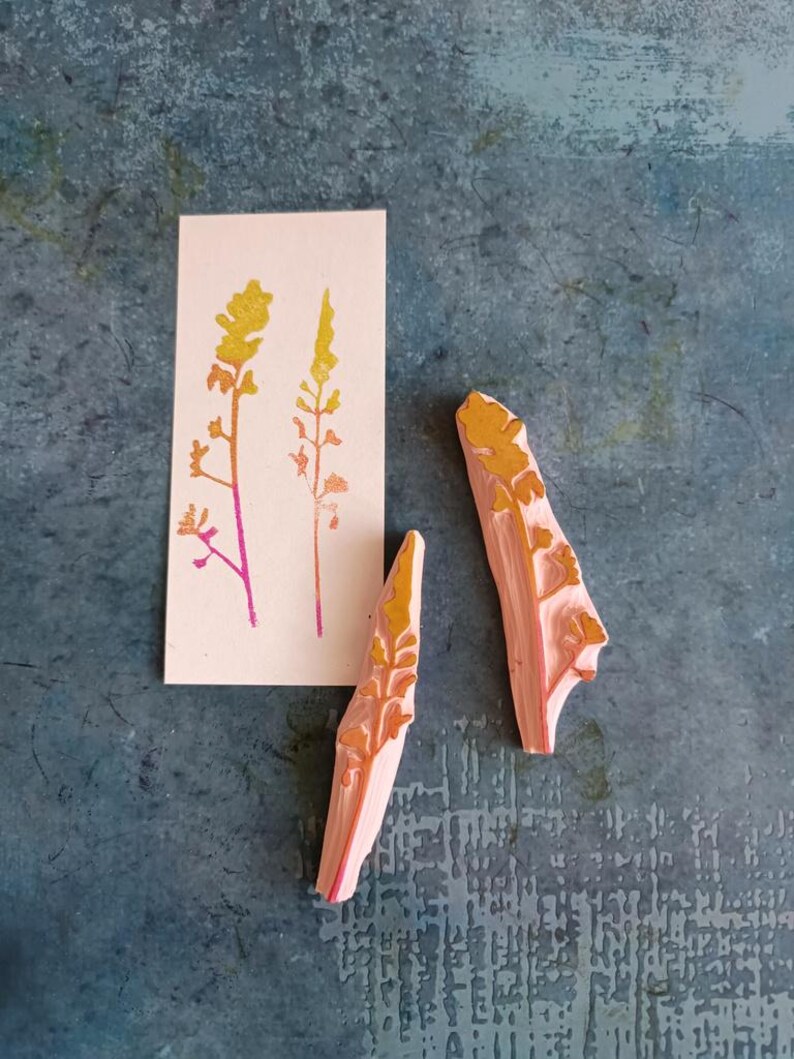 May include: Two pink rubber stamps with a yellow and orange floral design. The stamps are on a blue textured surface. A card with a yellow and orange floral design is in the background.