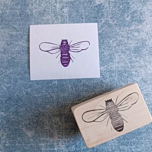 May include: A wooden stamp with a bee design next to a purple bee print on white paper. The bee stamp is rectangular and features a detailed outline of a bee. The bee print is a solid purple color.