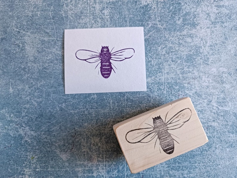 May include: A wooden stamp with a bee design next to a purple bee print on white paper. The bee stamp is rectangular and features a detailed outline of a bee. The bee print is a solid purple color.