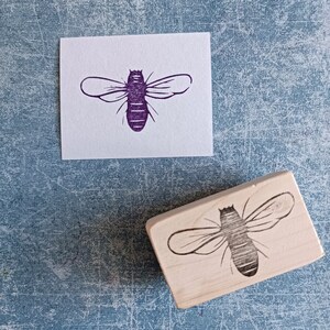 May include: A wooden bee stamp next to a purple bee print on white paper. The stamp has a rectangular wooden base with a detailed bee design. The bee print is a detailed illustration of a bee.
