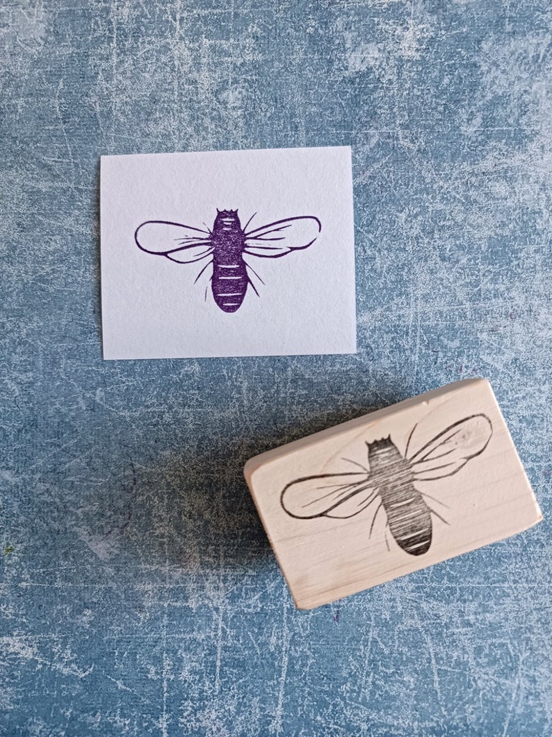 May include: A wooden bee stamp next to a purple bee print on white paper. The stamp has a rectangular wooden base with a detailed bee design. The bee print is a detailed illustration of a bee.