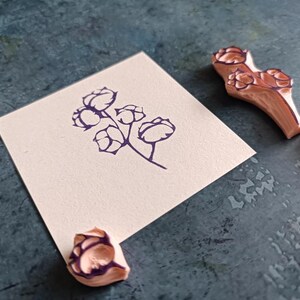 May include: A sprig of cotton flower stamped in dark purple ink on a square of off-white paper. Three wooden rubber stamps with cotton flower designs are next to the paper on a textured, teal surface. The stamps are light brown with dark purple details.