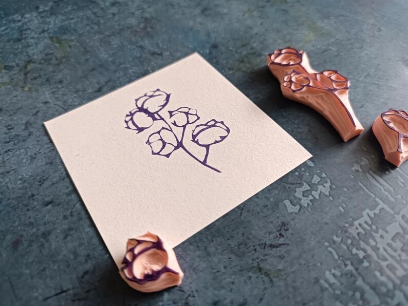 May include: A sprig of cotton flower stamped in dark purple ink on a square of off-white paper. Three wooden rubber stamps with cotton flower designs are next to the paper on a textured, teal surface. The stamps are light brown with dark purple details.
