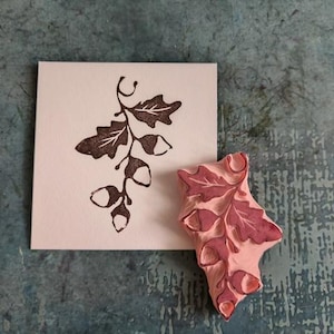 Puede incluir: Una impresión en tinta marrón de una rama de hojas y bellotas de roble sobre papel blanco, junto a un sello de goma rosa con el mismo diseño. El sello presenta un patrón detallado de hojas y bellotas, ideal para manualidades.