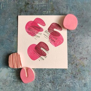 May include: A square card with three abstract pink shapes with black lines, and three pink rubber stamps. The stamps are in different shapes, including a circle, a curved shape, and a rectangular shape with lines. The background is a textured blue surface.