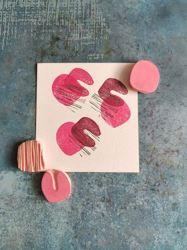 May include: A square card with three abstract pink shapes with black lines, and three pink rubber stamps. The stamps are in different shapes, including a circle, a curved shape, and a rectangular shape with lines. The background is a textured blue surface.