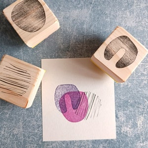 May include: Wooden stamps with abstract designs, including a circle, a U-shape, and parallel lines. A small square of paper displays a stamped design in purple and pink, with black lines. The stamps are on a textured blue surface.