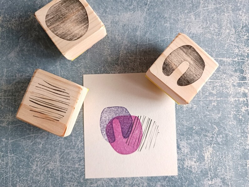 May include: Wooden stamps with abstract designs, including a circle, a U-shape, and parallel lines. A small square of paper displays a stamped design in purple and pink, with black lines. The stamps are on a textured blue surface.