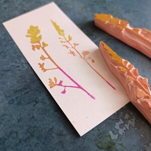 May include: Two wooden stamps with raised designs of stylised plants. The stamps are carved from light pink wood and have a yellow-orange colour on the raised design. The stamps are shown on a light pink card with two impressions of the plant design in yellow-orange and pink.