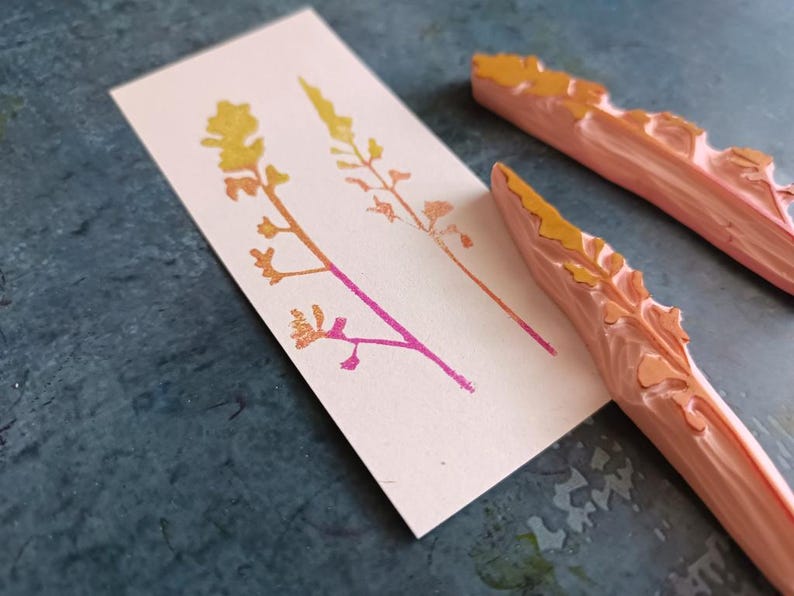May include: Two wooden stamps with raised designs of stylised plants. The stamps are carved from light pink wood and have a yellow-orange colour on the raised design. The stamps are shown on a light pink card with two impressions of the plant design in yellow-orange and pink.