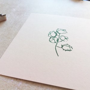 May include: A green botanical stamp impression of a plant with multiple seed pods on a white card. Two stamps are visible in the upper left corner. The card is on a light-colored surface.