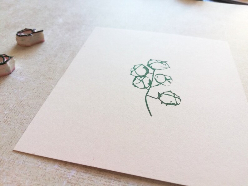 May include: A green botanical stamp impression of a plant with multiple seed pods on a white card. Two stamps are visible in the upper left corner. The card is on a light-colored surface.