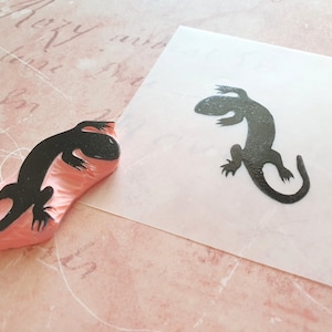 May include: A black lizard stamp next to a stamped image of the same lizard. The stamp is made of pink rubber and the lizard image is on a white surface. The background is a light pink paper with script.