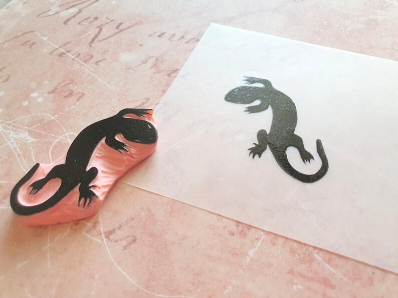 May include: A black lizard stamp next to a stamped image of the same lizard. The stamp is made of pink rubber and the lizard image is on a white surface. The background is a light pink paper with script.