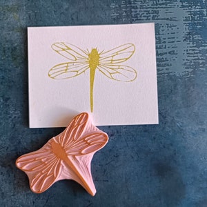 Könnte beinhalten: Ein rosa Stempel aus Gummi mit einer Libelle mit detaillierten Flügeln. Der Stempel befindet sich auf einem blauen Hintergrund neben einer weißen Karte mit einer goldenen Libellenillustration.