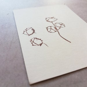 May include: A brown floral stamp impression on off-white paper. The design features stylized flower buds on stems. The stamp is visible in the corner, with a pink and black design. The paper is on a gray surface.