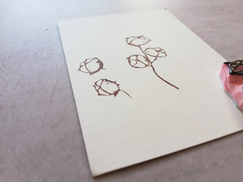 May include: A brown floral stamp impression on off-white paper. The design features stylized flower buds on stems. The stamp is visible in the corner, with a pink and black design. The paper is on a gray surface.