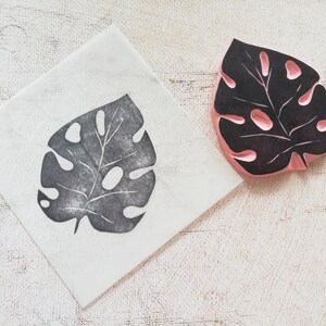 May include: A black and pink rubber stamp of a Monstera leaf, next to a print of the same leaf on a white square. The stamp has a detailed leaf design with cut-out holes. The print is in dark gray ink.