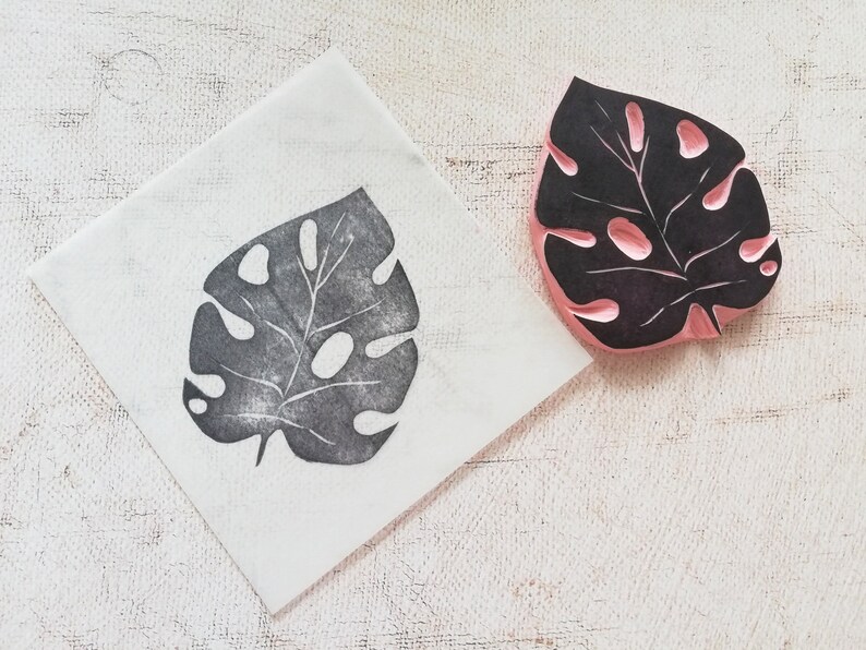 May include: A black and pink rubber stamp of a Monstera leaf, next to a print of the same leaf on a white square. The stamp has a detailed leaf design with cut-out holes. The print is in dark gray ink.