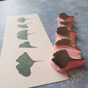 May include: A set of six ginkgo leaf stamps, with a pink rubber base and black ink. The stamps are arranged in a row, with the largest leaf at the top and the smallest leaf at the bottom.